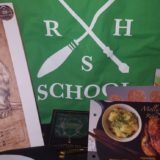 Geek Gear July 2018: Slytherin tote bag showing a wand and broom crossed, with an initial of each House in the blank space between the wand and broom. Words on the tote say: Wizarding School