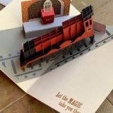 Pop-Up Hogwarts Express Card from Insight Editions showing the red steam engine car with a school trunk and Hedwig’s cage waiting on the platform