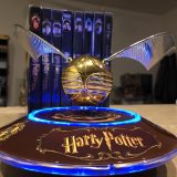 Harry Potter GOLDEN SNITCH™, rotating on base in the light, with “Potter” movie cases in the background