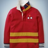 The Mini Boden Hogwarts Rugby Shirt in red is collared with cuffs and features horizontal gold stripes near the hem and a Harry Potter glasses-and-scar design in the front pocket area. It retails at £30.