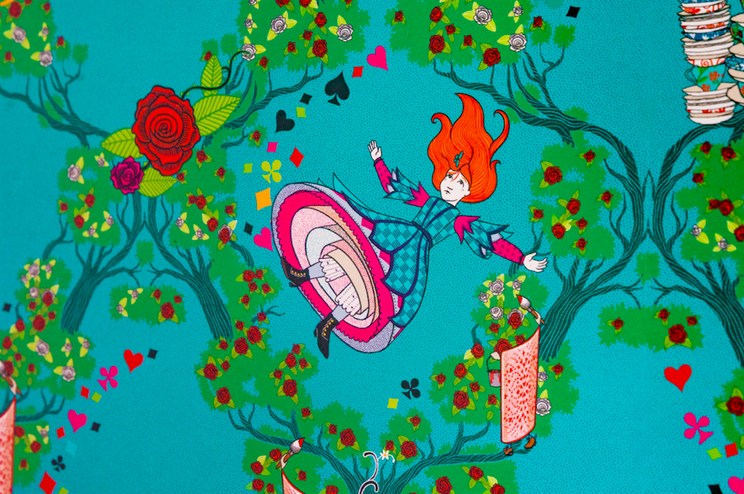 This MinaLima “Alice’s Adventures in Wonderland” wallpaper features Alice as she falls through the rabbit hole to Wonderland surrounded by white and red roses on a light blue background.